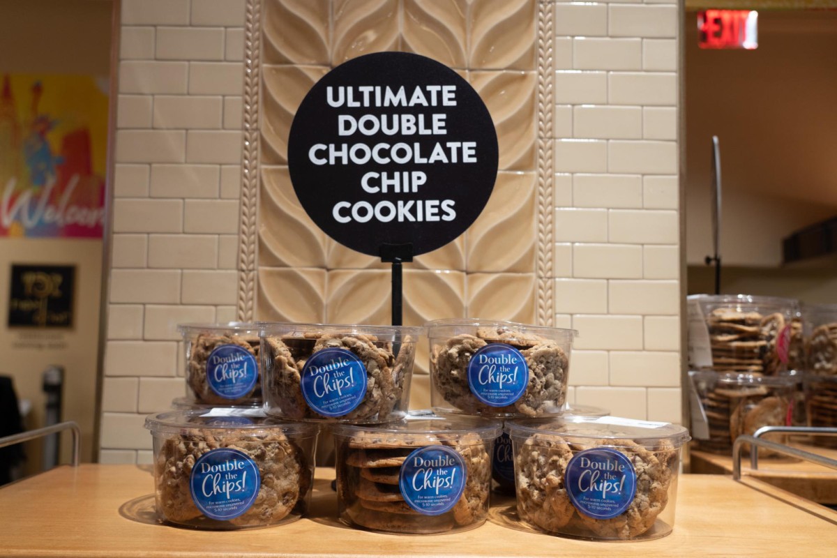 Six buckets of chocolate chip cookies stacked on a wooden shelf. Behind them is a black sign that says “ULTIMATE DOUBLE CHOCOLATE CHIP COOKIES” in white letters.