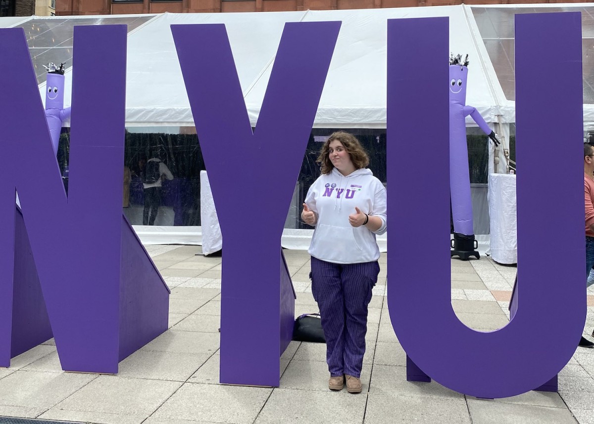 A+student+is+standing+between+giant+purple+letters+that+spell+out+N.Y.U.+She+is+wearing+a+white+and+purple+N.Y.U.+hoodie.