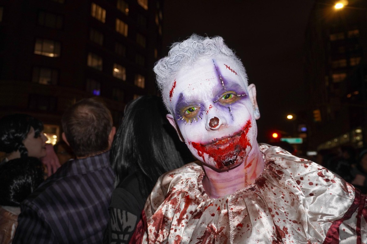 A man dressed in a bloody clown costume with white face paint and hair. He has purple eyeshadow, green eye contacts and a bloody mouth.