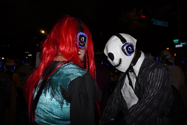 A person with a red wig and a blue-and-black shirt. Another person in a ghost mask and a black-and-white suit. They are both wearing headphones.