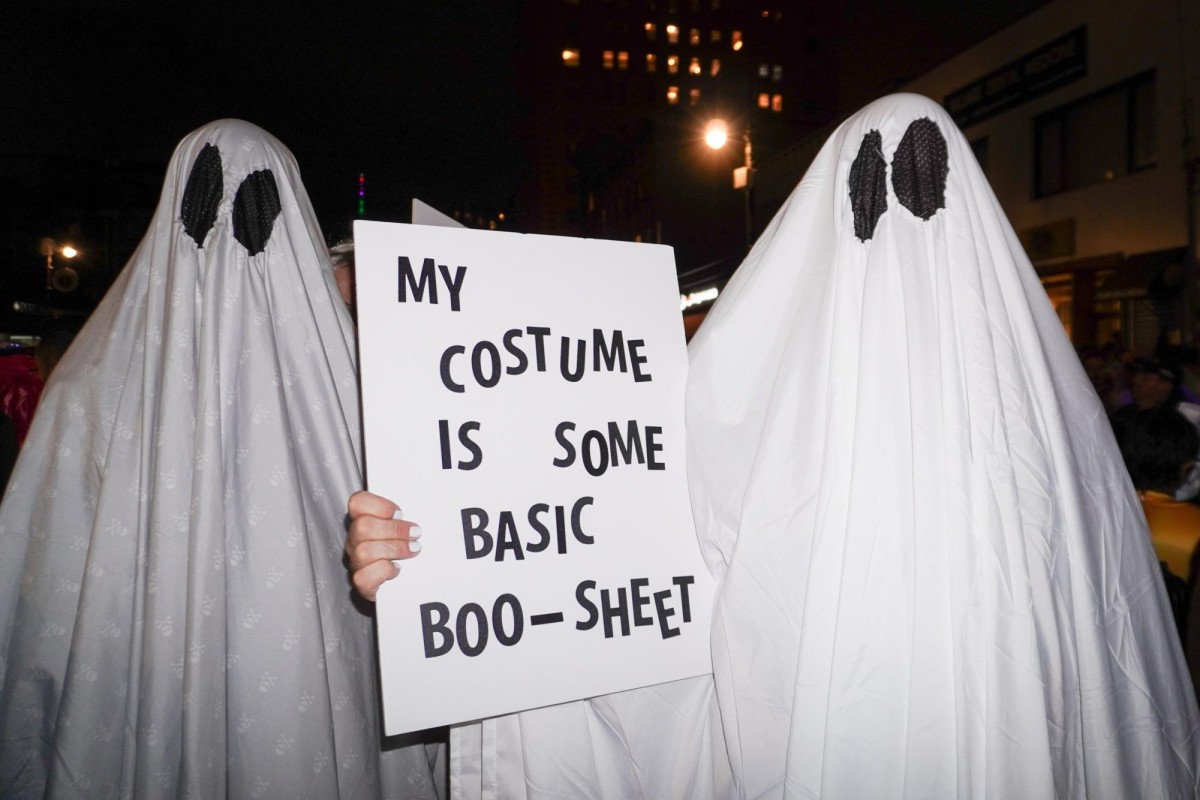 Two figures covered with white sheets standing side by side. The figure on the right holds up a white sign with black letters that read “MY COSTUME IS SOME BASIC BOO-SHEET.”