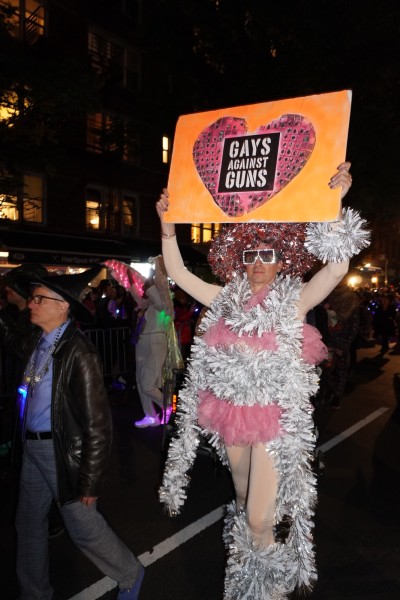 A man wearing a silver-and-pink skirt, a red-and-silver wig, and a pair of silver glasses holding up an orange sign with a pink heart on it. At the center of the heart is a square with “GAYS AGAINST GUNS” written on it.