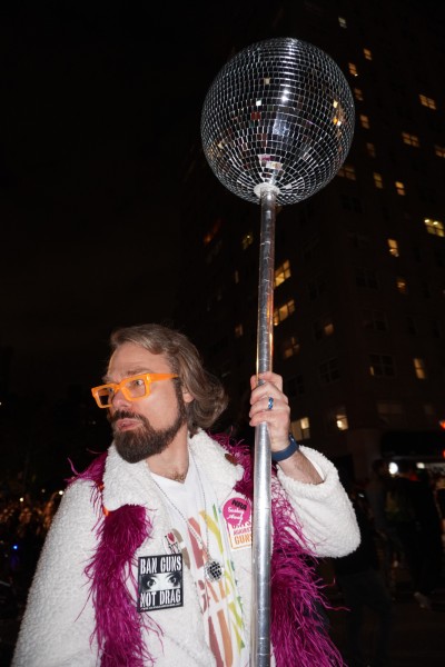 A man with brown hair, a beard and a pair of orange glasses wearing a white coat and a fuschia scarf holds a metal staff with a disco ball on the top. He is wearing pins that read "BAN GUNS NOT DRAG" and "NRA Sashay Away."