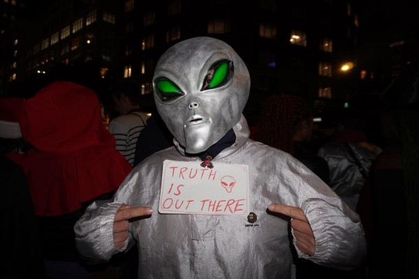 A person in a gray alien costume with a paper sign attached to his collar that has the text “TRUTH IS OUT THERE.”