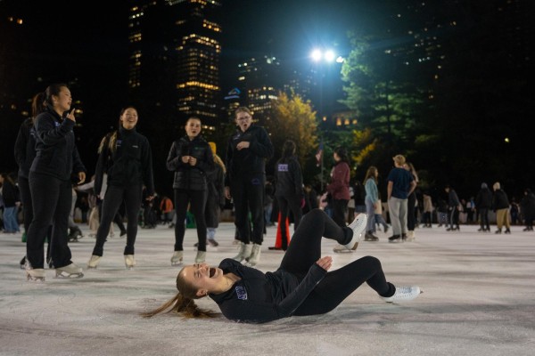 A person lays on the ice in a black long sleeve with the letters N.Y.U. embroidered on the front in purple, black pants and white ice skates. Four N.Y.U. Figure Skating members all wearing black outfits watch in the background with shocked expressions on their faces.