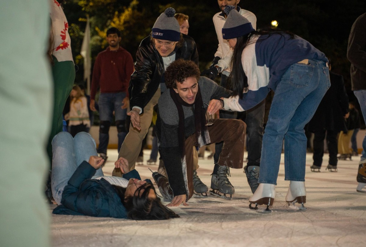A man in a grey and black sweater and brown trousers kneels on the ice, while a woman wearing a blue jacket and blue jeans lies beside him. A group of people in various colored outfits surrounds them and try to help them up.