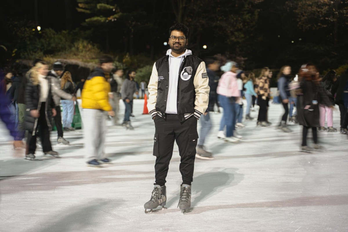 A person in a white hoodie, black and white varsity jacket, black pants and gray ice skates stands for a portrait on the ice rink.