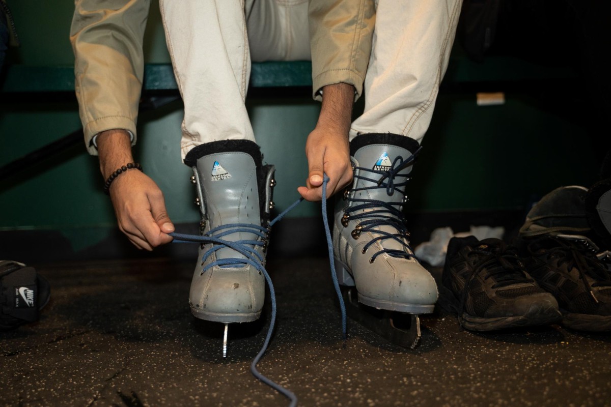 A person in a beige top and white pants laces up gray ice skates.