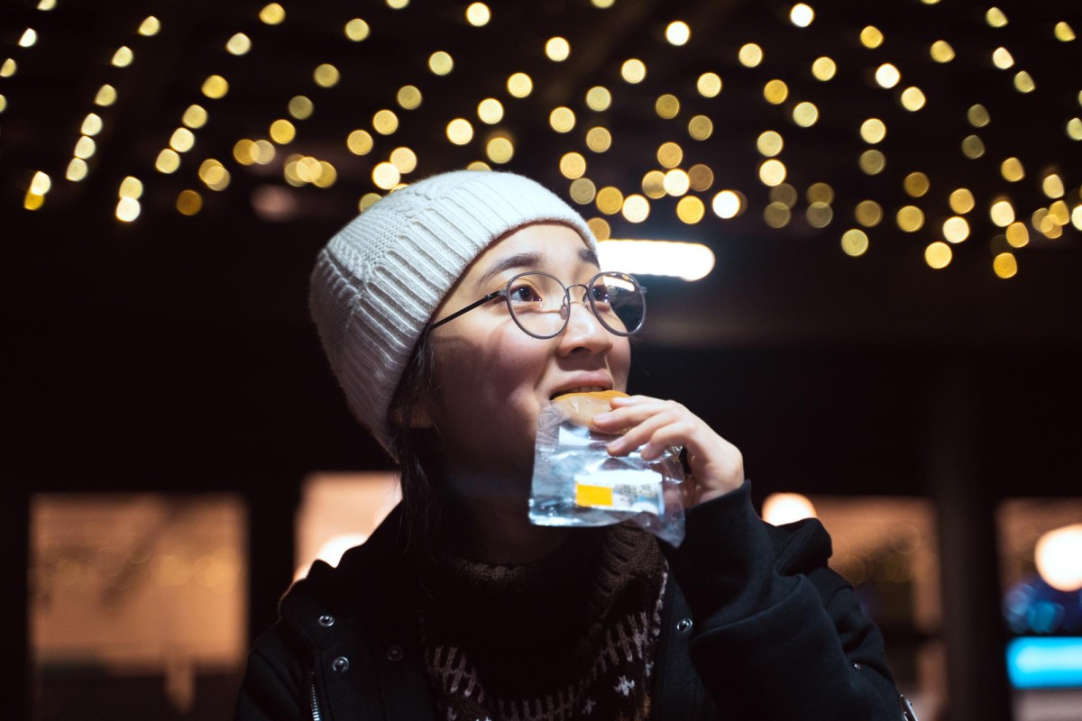 A person in a white beanie, round glasses, black overcoat and dark sweater eats food.