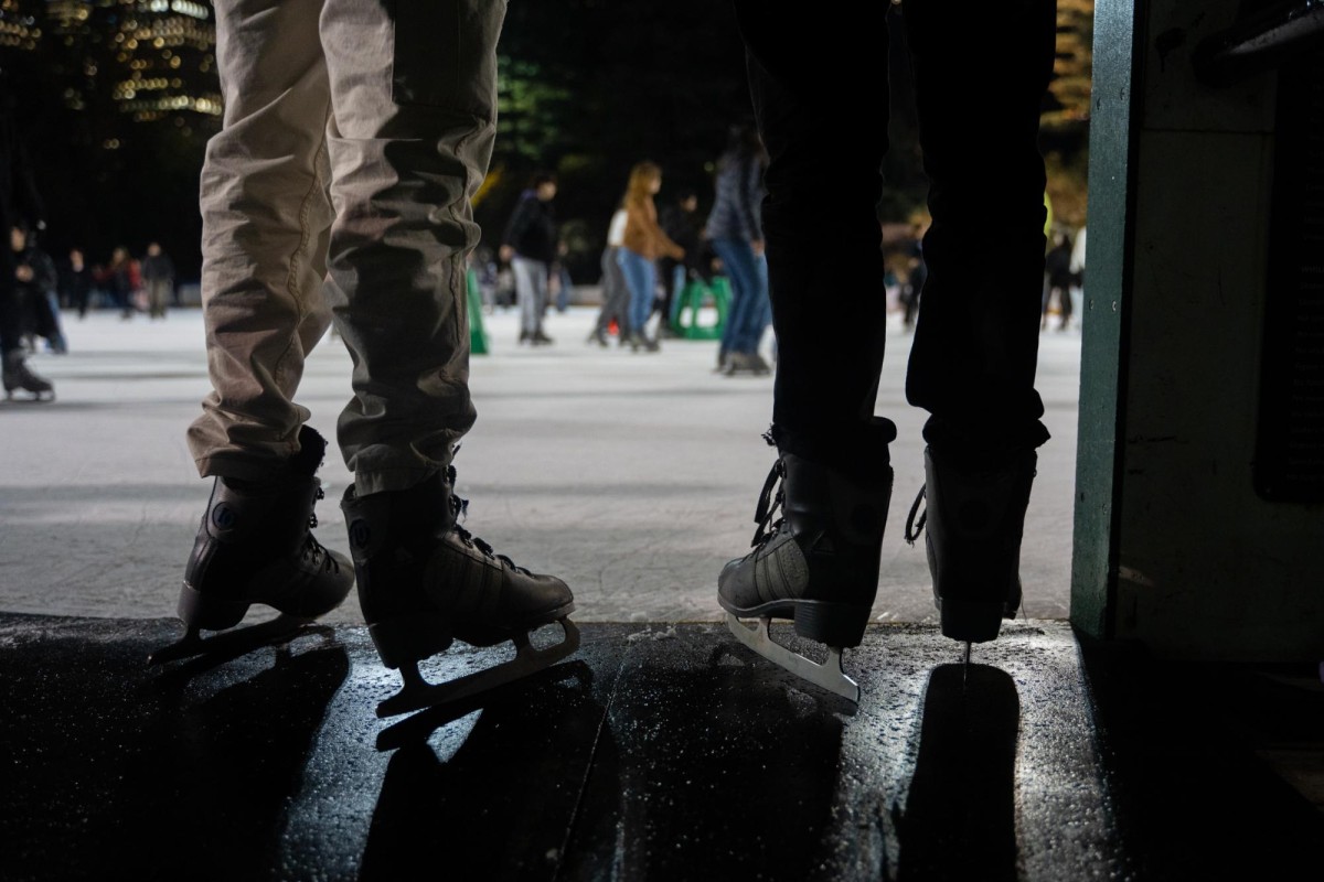 Two people enter the ice rink with gray ice skates on, one with beige pants and the other with black pants.