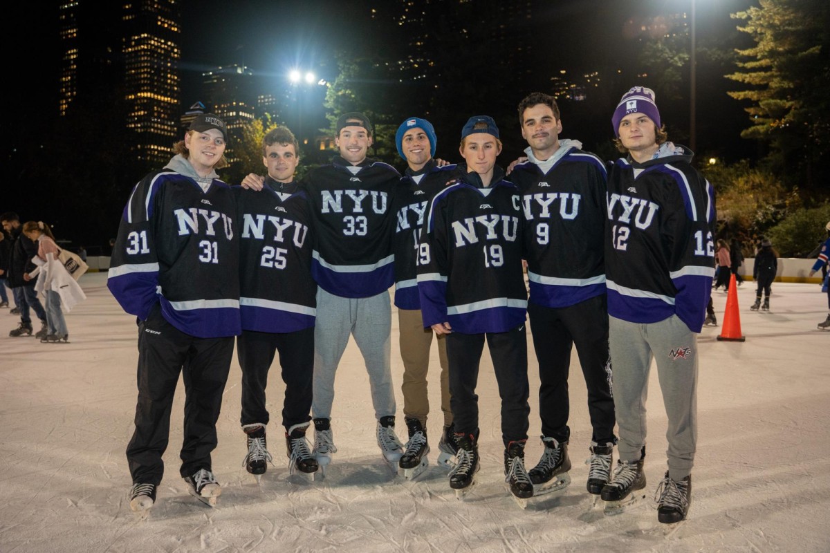 Members of the N.Y.U. Hockey team in black and purple accented hockey jerseys and various colored pants, all wearing ice skates stand on the ice rink.