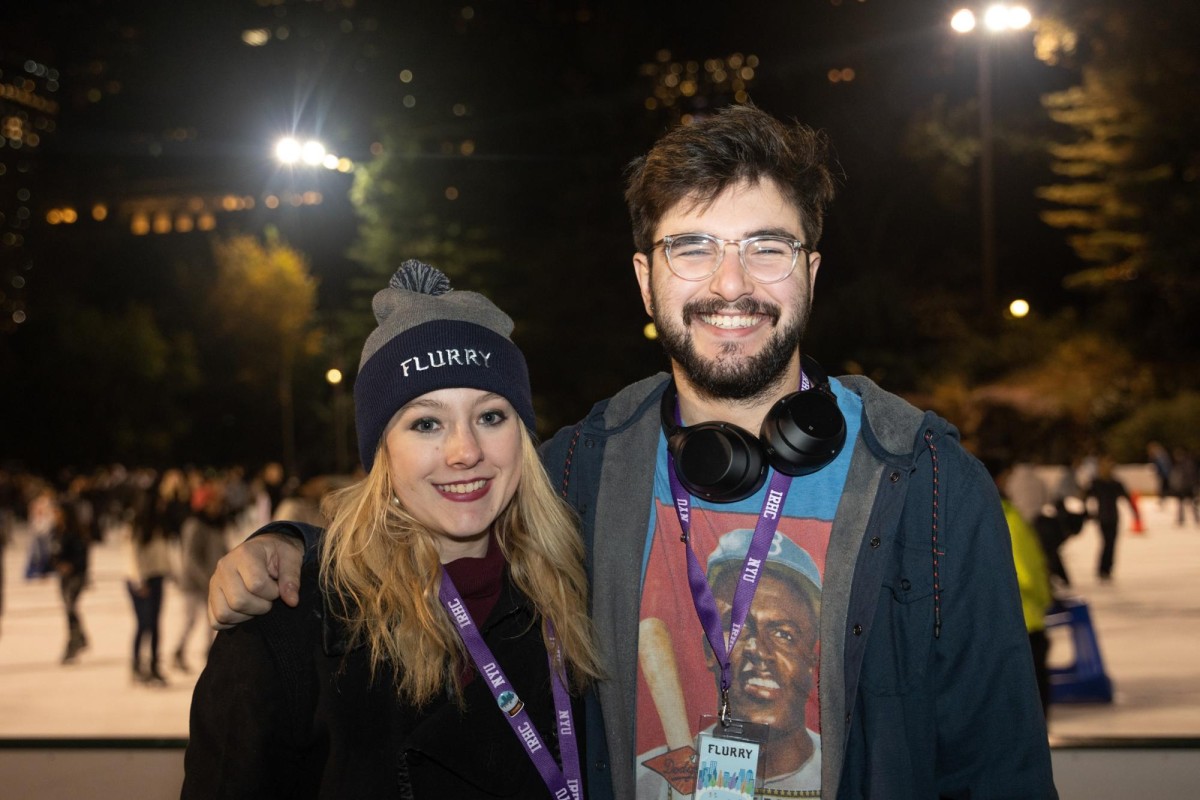 A woman in a gray and navy beanie with blond hair and a black jacket stands next to a man with black hair and a beard wearing clear glasses, black headphones, a navy jacket and a blue t-shirt with a graphic of a baseball player. Both wear purple N.Y.U. I.R.H.C. lanyards.