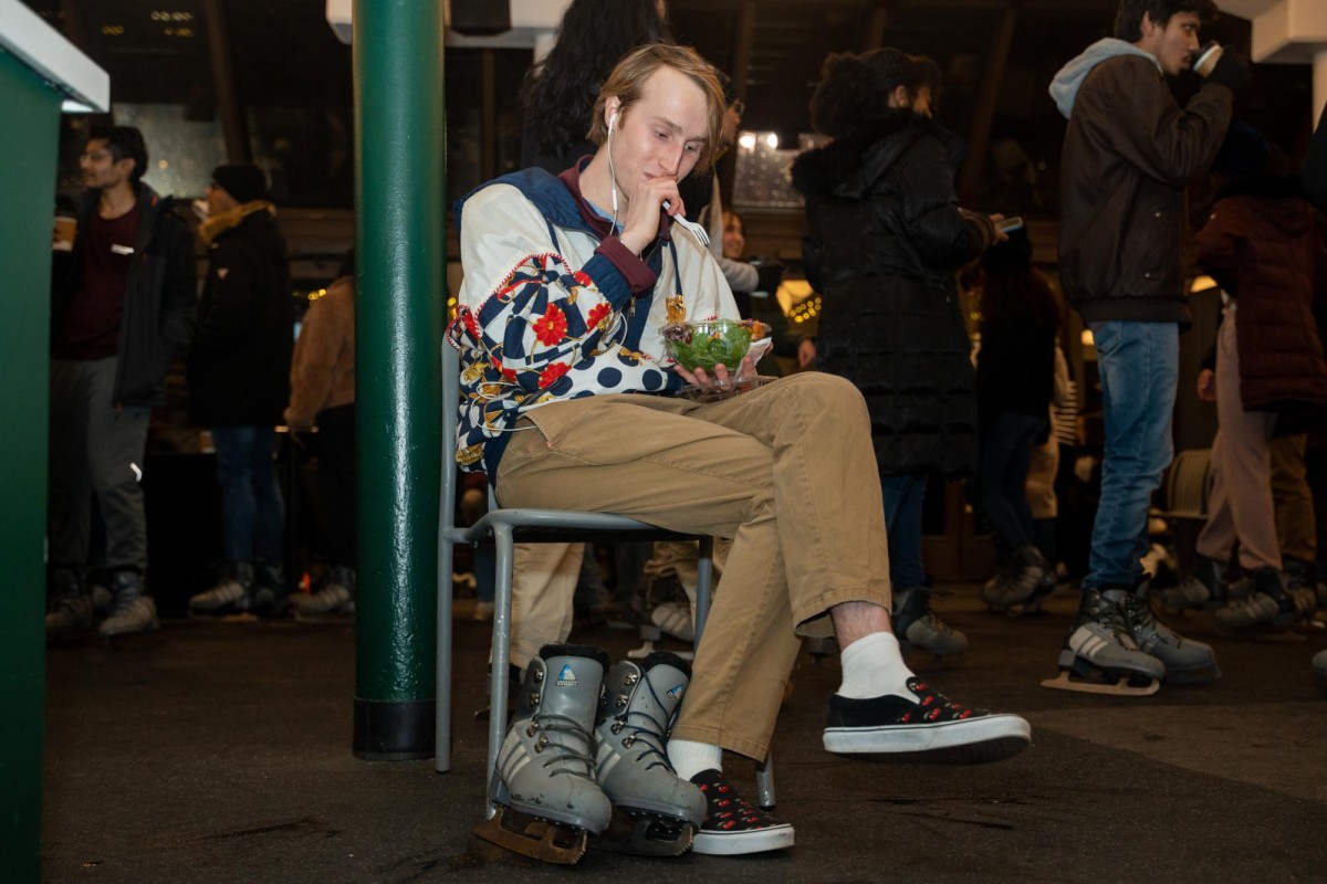 A person with blonde hair sits eating a salad in a white jacket with navy blue and red accents, beige pants and white socks with black slip-on shoes.