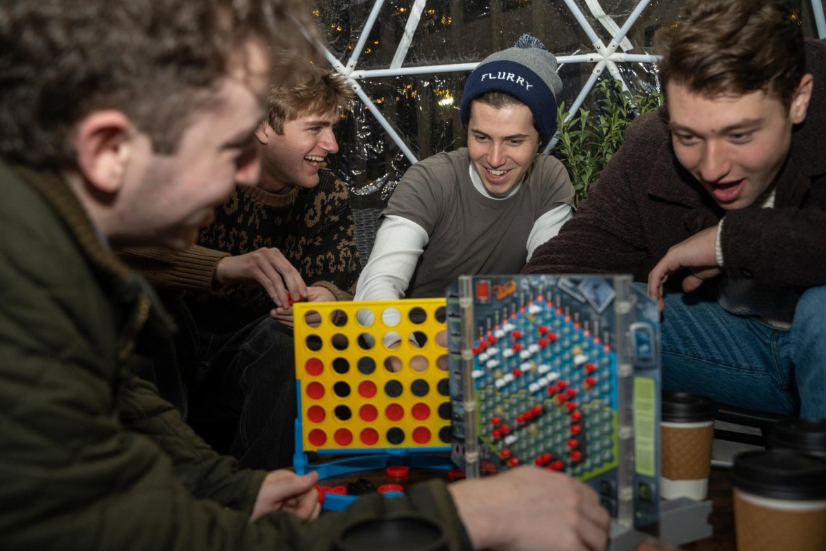 Four smiling people play Connect 4 and Battleship. From left to right, a person wears a green jacket, a beige and brown sweater, a gray t-shirt and white long sleeve undershirt, and a brown jacket.