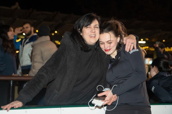 A person with black hair and in a black coat hugs an N.Y.U. Figure Skating member who is wearing a black long sleeve with the letters N.Y.U. embroidered on the front in purple as well as the member’s name embroidered on the left shoulder in white.