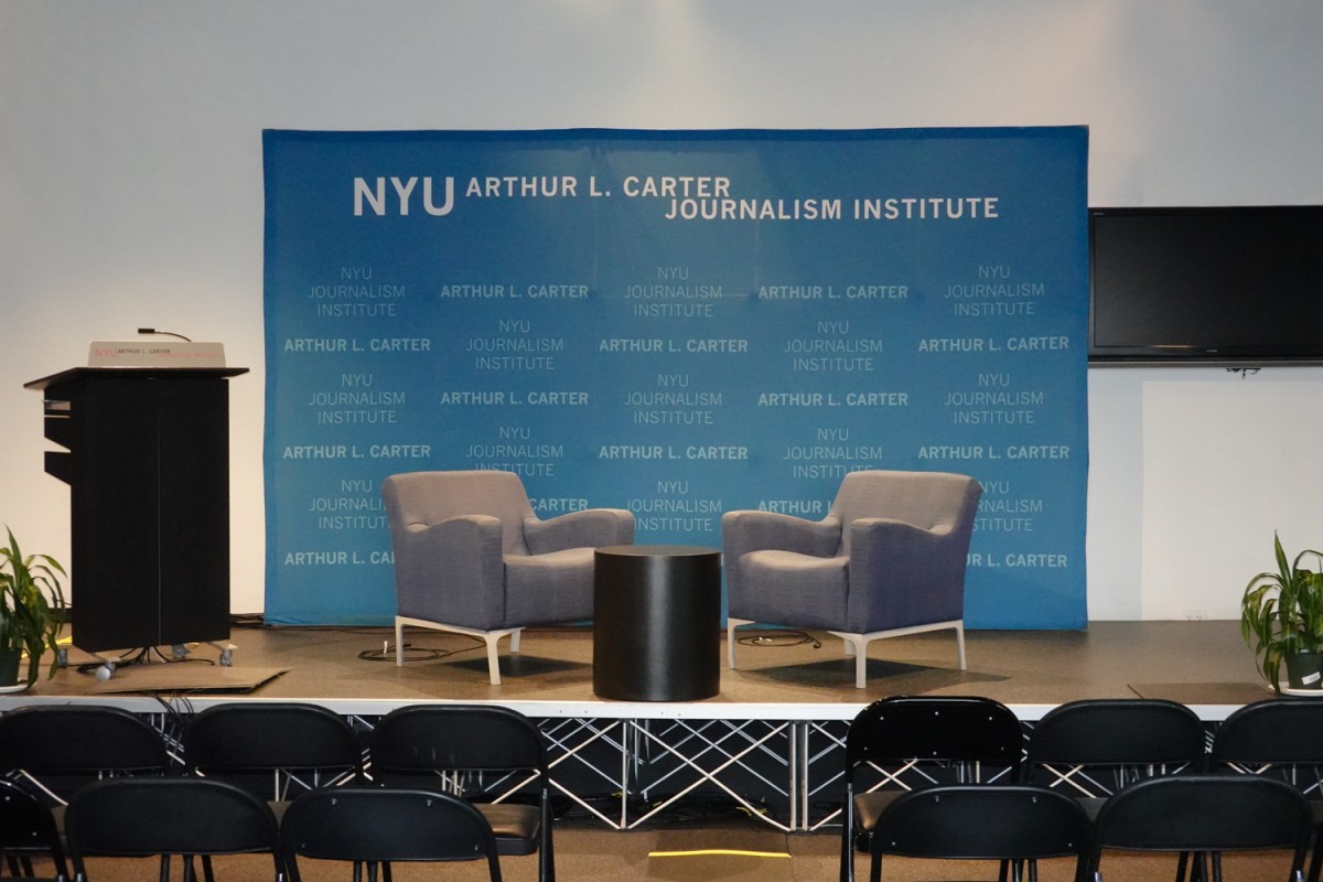 The front view of a stage with a black podium, two chairs, a small black, round table and a blue board that says “N.Y.U. ARTHUR L. CARTER JOURNALISM INSTITUTE” on it.