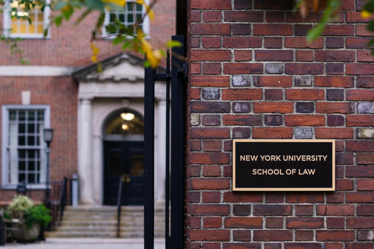 To the right there is a sign on a brick wall that reads, “New York University School of Law.” Behind that wall is the entrance to a brick building.