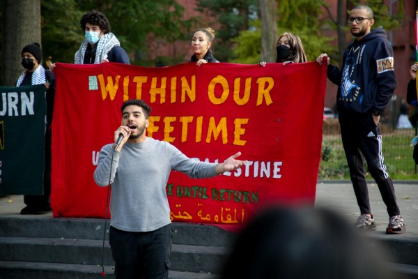 A man holds a mic and speaks in front of a red banner that says “WITHIN OUR LIFETIME.”