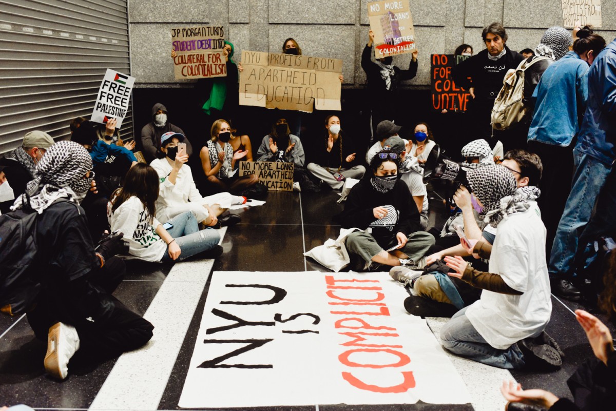 A group of students, both sitting and standing around a large white banner which reads “N.Y.U. is Complicit.”