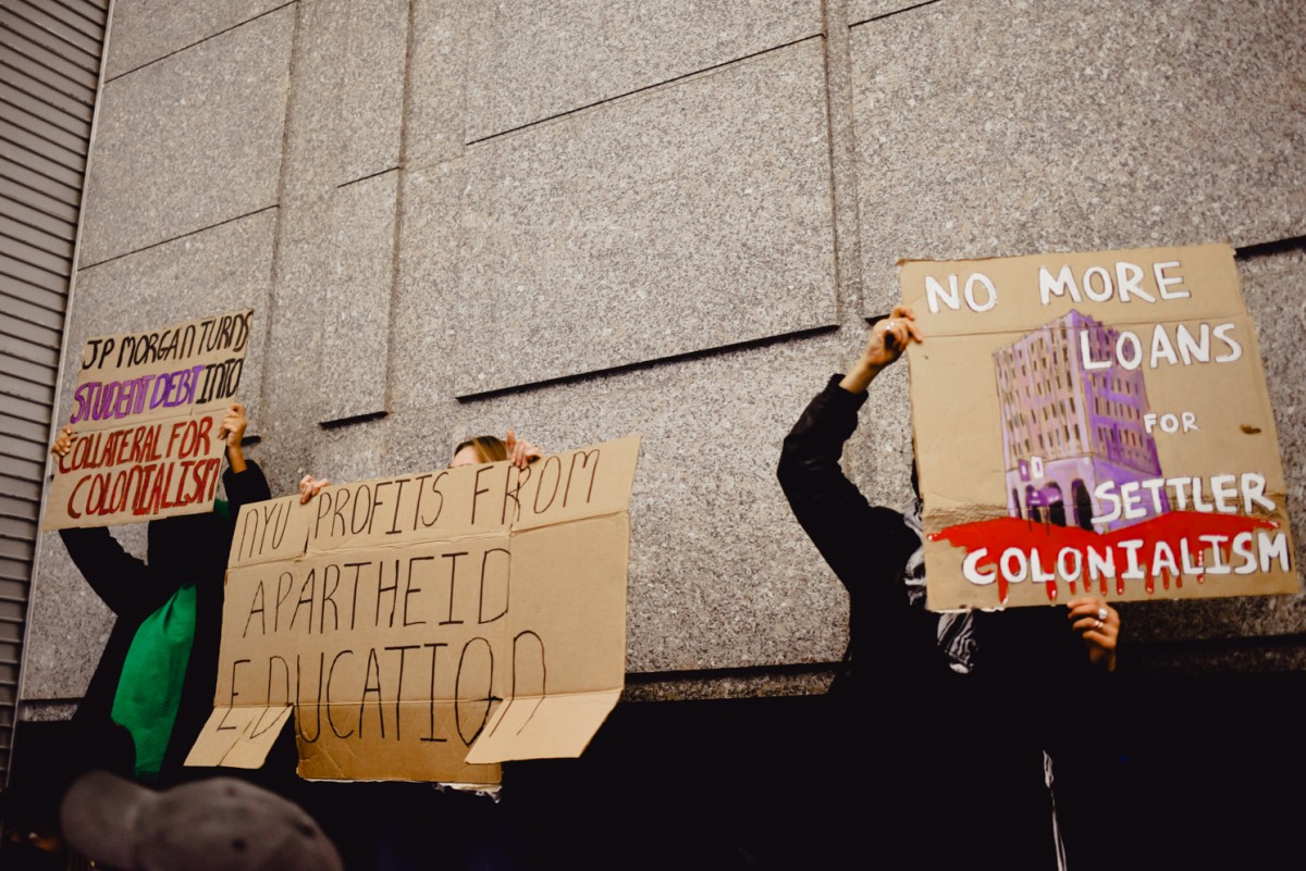 Three people holding cardboard signs over their faces which read “J.P. MORGAN TURNS STUDENT DEBT INTO COLLATERAL FOR COLONIALISM,” “N.Y.U. PROFITS FROM APARTHEID EDUCATION,” and “NO MORE LOANS FOR SETTLER COLONIALISM.”
