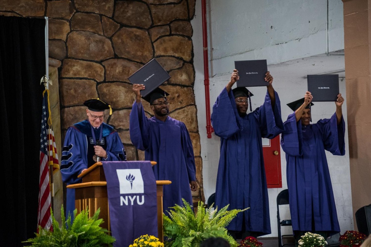 Three+people+wearing+purple+graduation+gowns+and+black+graduation+caps+are+standing+raising+diplomas.+To+the+left+is+former+N.Y.U.+president+standing+behind+a+purple-and-white+N.Y.U.+sign.
