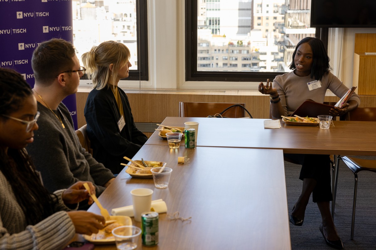 A wooden, L-shaped table with four people sitting with paper plates of food in front of them. Three of them are on one side of the table, looking at the one person on the other side. To the left there is a purple sign that reads N.Y.U. Tisch.