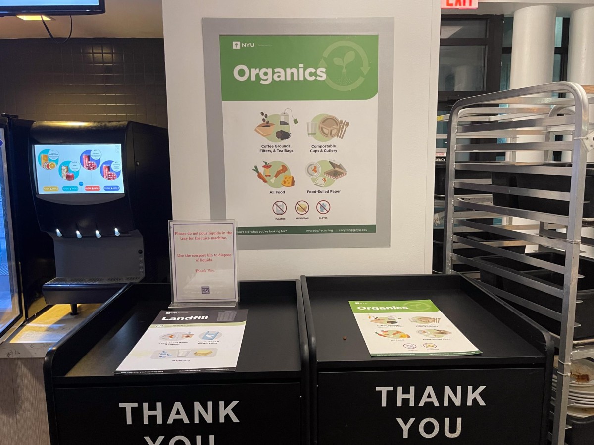 A white and green sign on a white wall. It says, “Organics,” and has images of food items and utensils on it. Beneath it, there are two black garbage bins that have “Thank You,” written on them in white letters.