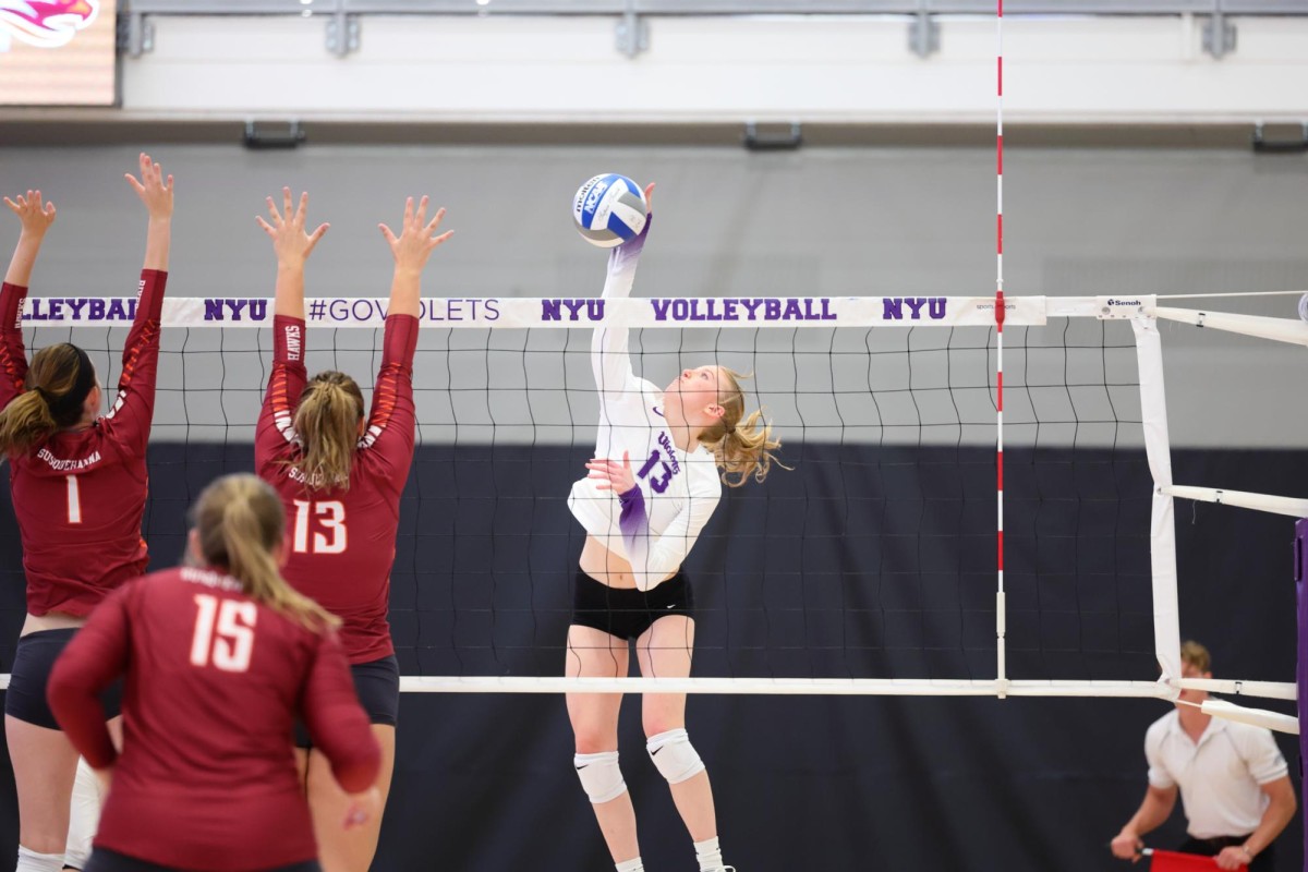 A girl in a white jersey that says “Violets” and the number 13 is jumping and hitting a volleyball over the net. Two women from the opposing team with red jerseys are jumping and trying to block the ball on the other side of the net.