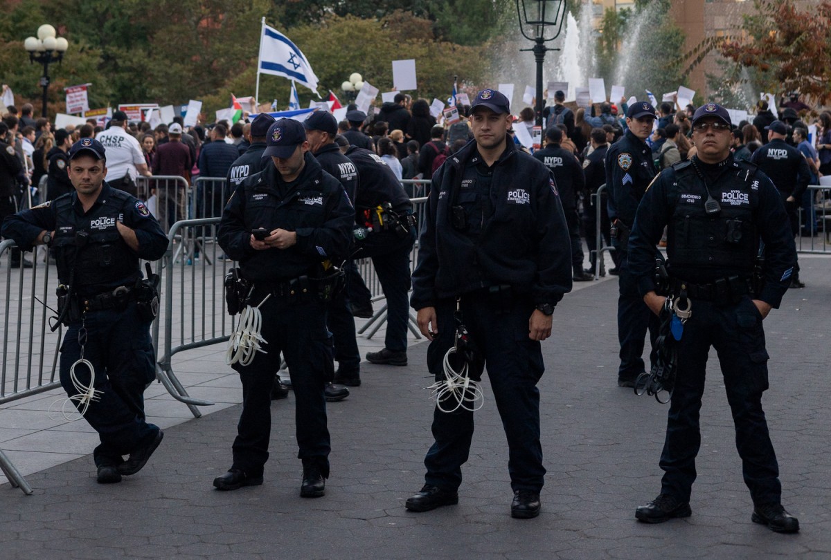 Four police officers standing in a line in Washington Square Park. One of the officers is leaning on a metal bar fence. Behind them are pro-Palestinian and pro-Israeli protesters congregated around a fountain.
