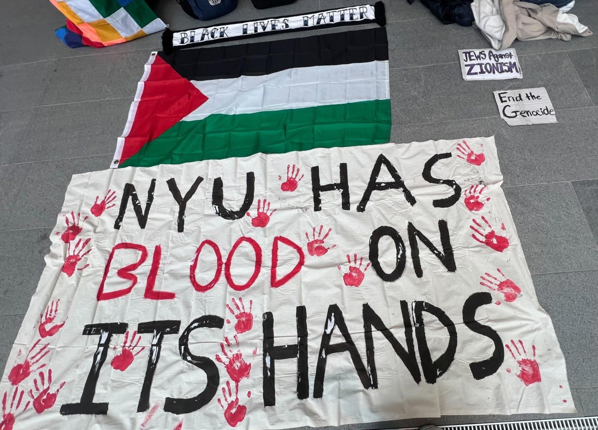 A white banner with the words "N.Y.U. HAS BLOOD ON ITS HANDS" with the word "BLOOD" written in red. Surrounding the words are multiple red handprints. Above it is a Palestinian flag. They are laid on a grey tile floor next to small pieces of paper with slogans on them.