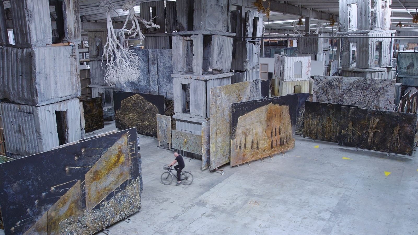 A wide shot of a warehouse-like space. The walls and the floor are all gray. There are some large paintings leaning on stacks of gray stacks of boxes and white trees hanging off of the ceiling. There is a man in a black T-shirt and black pants riding a bike.