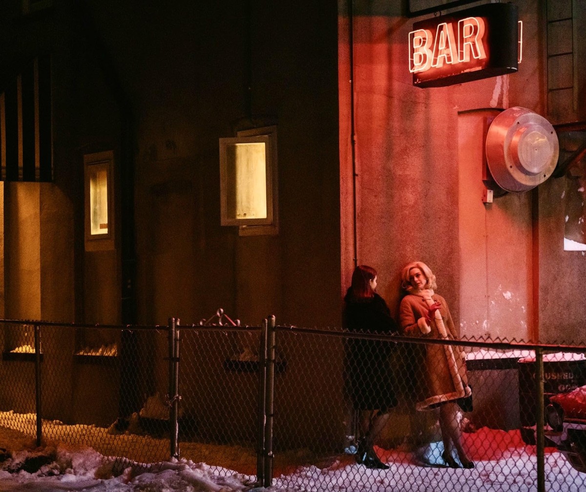 A person with blonde hair and a large beige overcoat leans against a concrete wall while conversing with a person standing next to them with brown hair and a black coat. Above them, a large neon red sign that reads “BAR” illuminates the snow on the ground in a red glow.