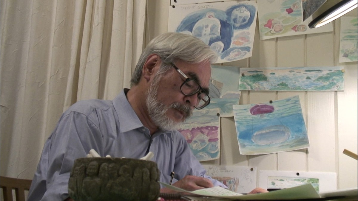 A man (Hayao Miyazaki) with a white beard, white hair and glasses is sitting down, looking at a piece of paper. Behind him, there are paintings hanging on the wall.