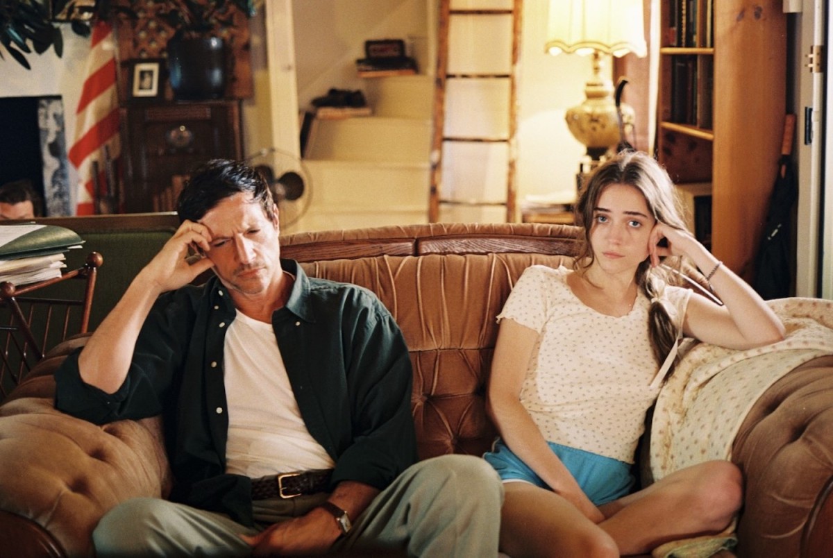 Two people are sitting on a brown couch. Both of them appear upset and are leaning their head on their hands. The man on the left has short dark hair and is wearing a black shirt. The girl on the right has long, brown hair and is wearing a white shirt with blue shorts.