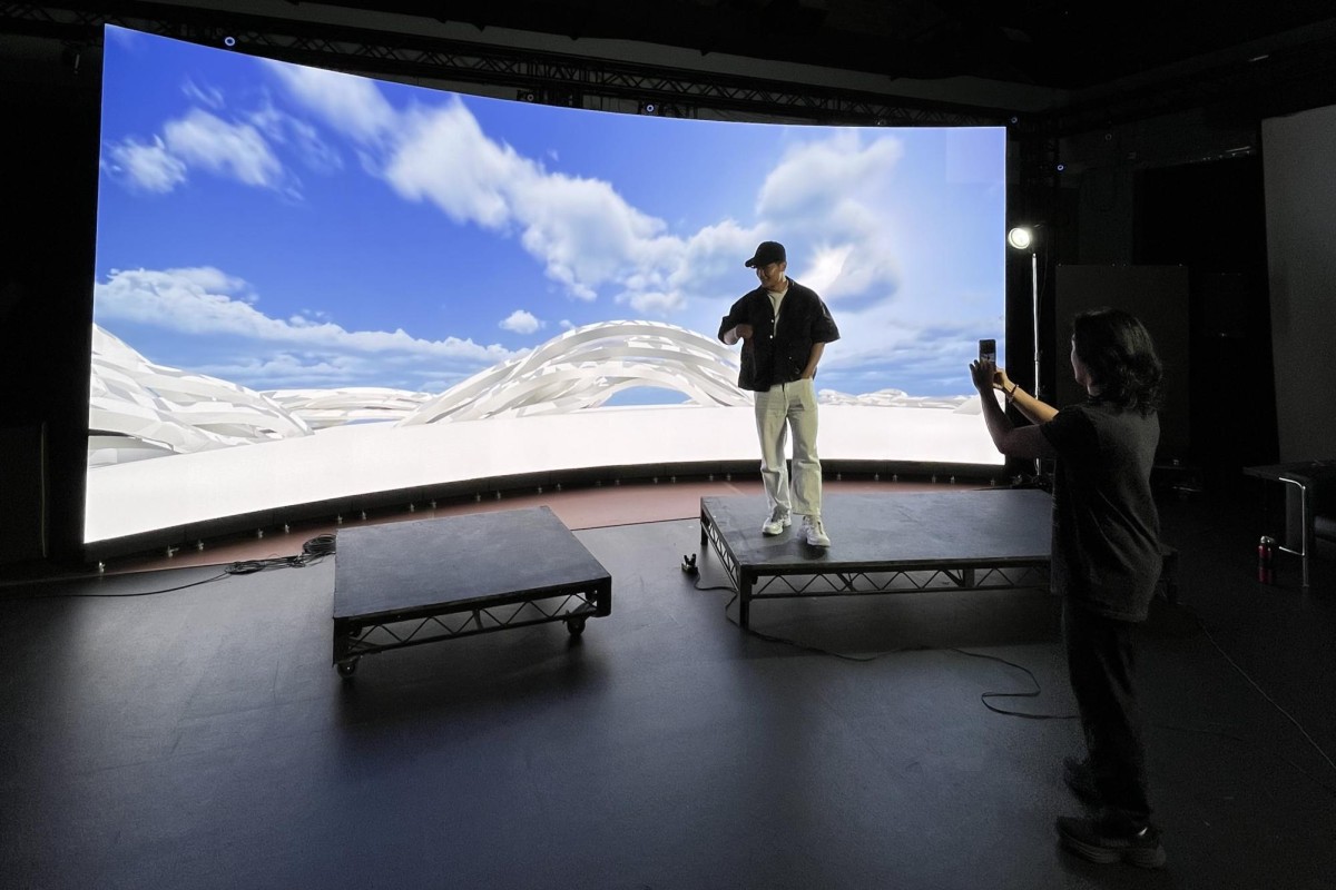 A person in a black hat and shirt with white pants and shoes stands in a dark room on a platform in front of a large curved digital screen with a projection of a blue sky with white clouds and white ground.