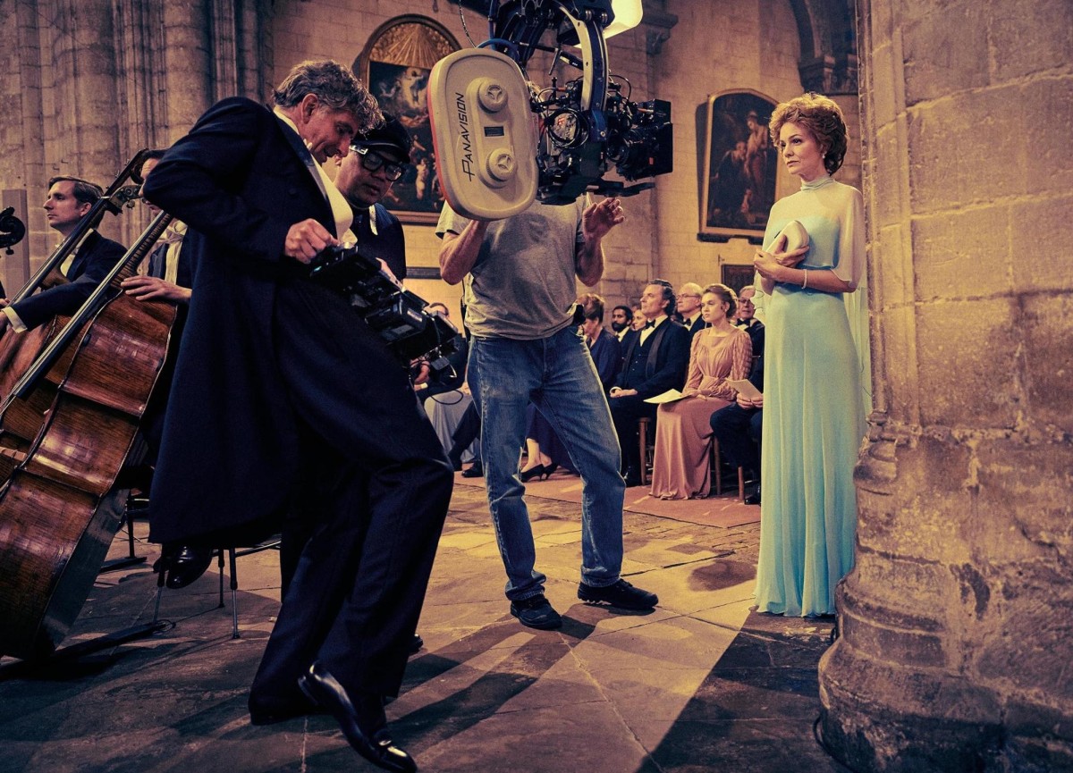 A+small+film+crew+stands+in+a+cathedral+with+camera+equipment%2C+filming+a+woman+in+a+long+blue+dress.+A+woman+in+a+long+blue+dress.+Behind+the+film+crew%2C+two+people+sit+with+cellos%2C+and+larger+group+of+people+are+seated+like+an+audience.