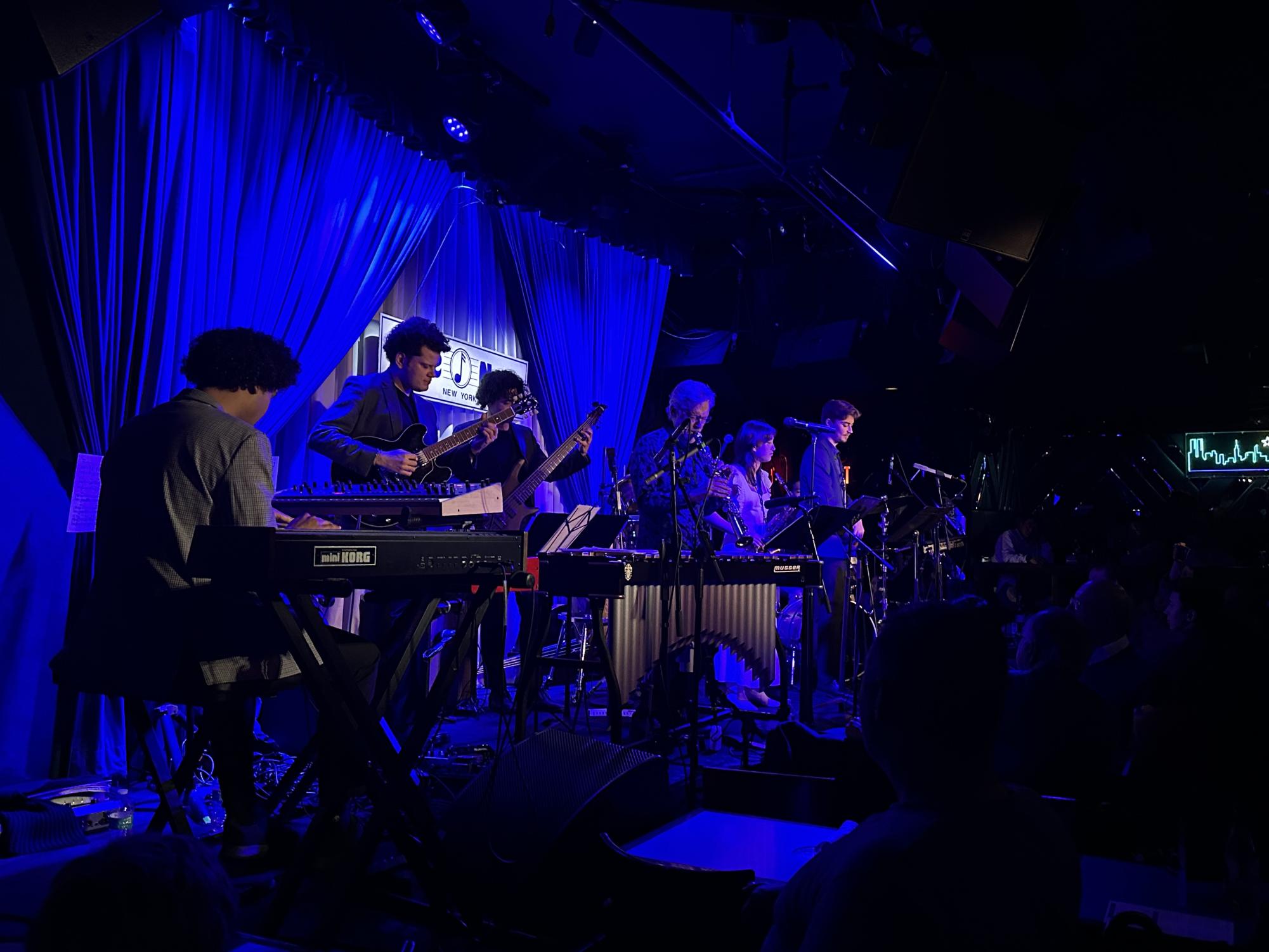 A jazz ensemble, consisting of six musicians with different instruments, performs on a stage. On the background of a stage is a curtain that is lit blue and a white sign with the name “Blue Note” on it.