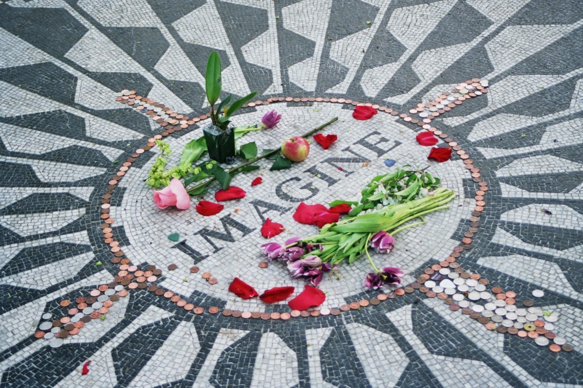 A pile of flowers, petals and pennies sitting on a mosaic circle made up of black-and-white tiles with the capitalized word “IMAGINE” in the center.