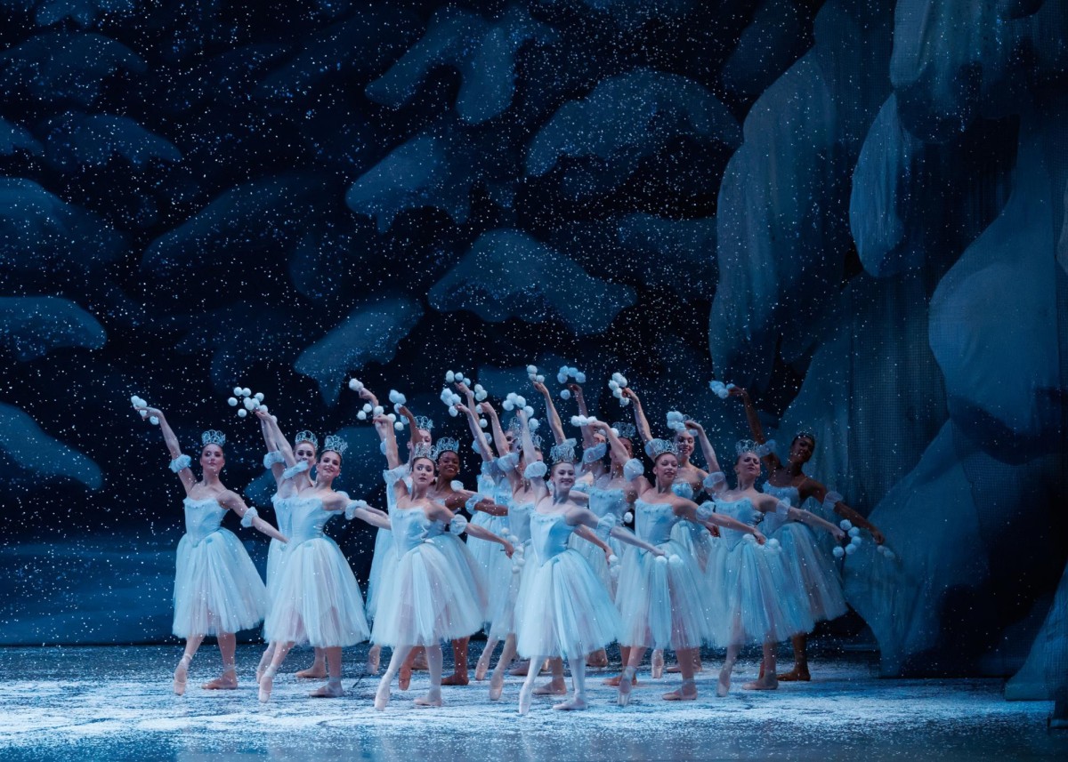 Fake snow falls around a group of ballet dancers dressed in light blue costumes who stand on a stage that is designed to look like a forest covered in snow.