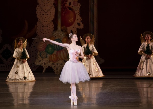 A ballet dancer wearing a light pink costume stands on pointe and holds a wand on stage. Three dancers stand at the back of the stage dressed as angels.