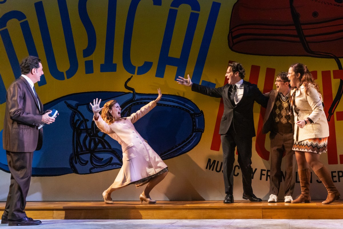 Reg Rogers in a suit with a blue tie, Katie Rose Clarke in a pink long coat with her arms outstretched, Jonathan Groff in a black suit with a black tie, Daniel Radcliffe in a brown suit and sweater and Lindsay Mendez wearing a white coat with a multi-colored striped dress standing on a stage.