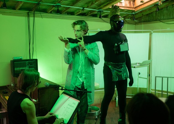 A person wearing a black bodysuit and face mask stretches their arm out, and a person standing next to them is holding their arm. A person sits to the left with a microphone and a music stand, reading a script that is on the stand.