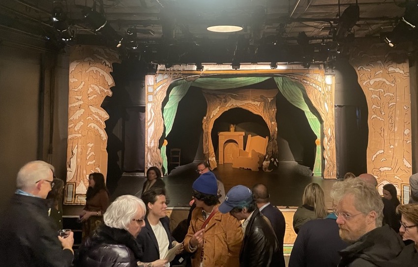 People standing in front of a stage with theater decor. There are brown cardboard that have trees and tree branches on them surrounding the stage.