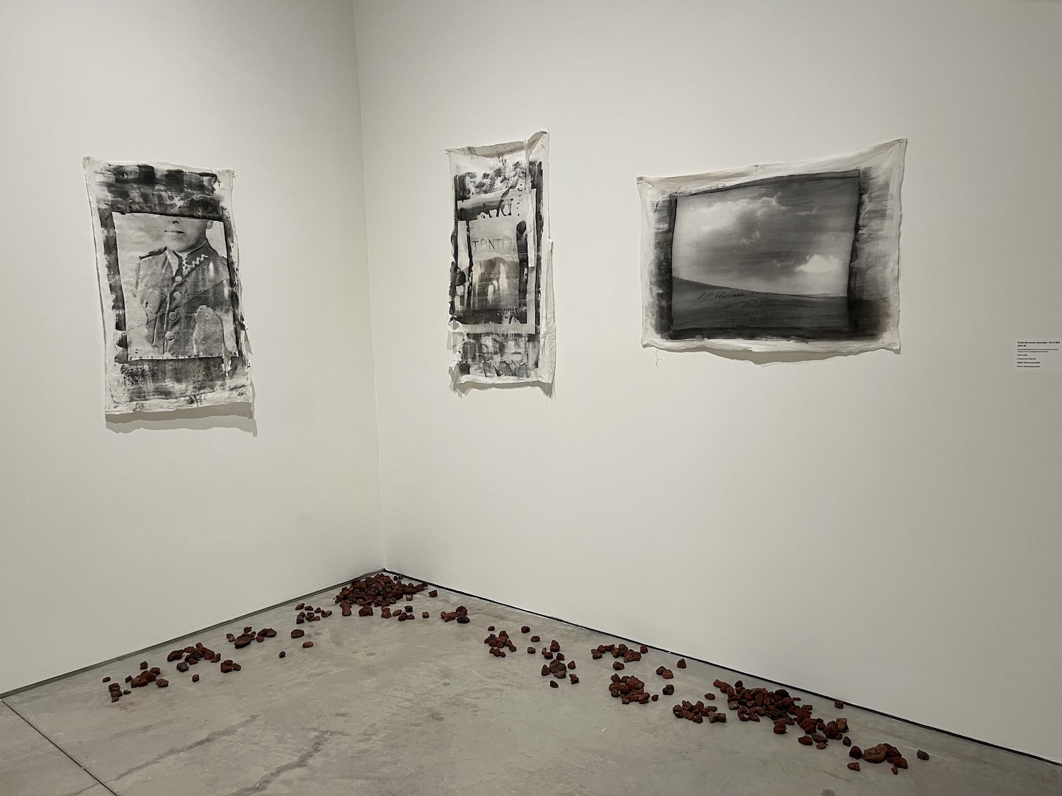 A corner of a room with white walls. There are three black and white paintings on fabric hung up. On the floor are small volcanic rocks.