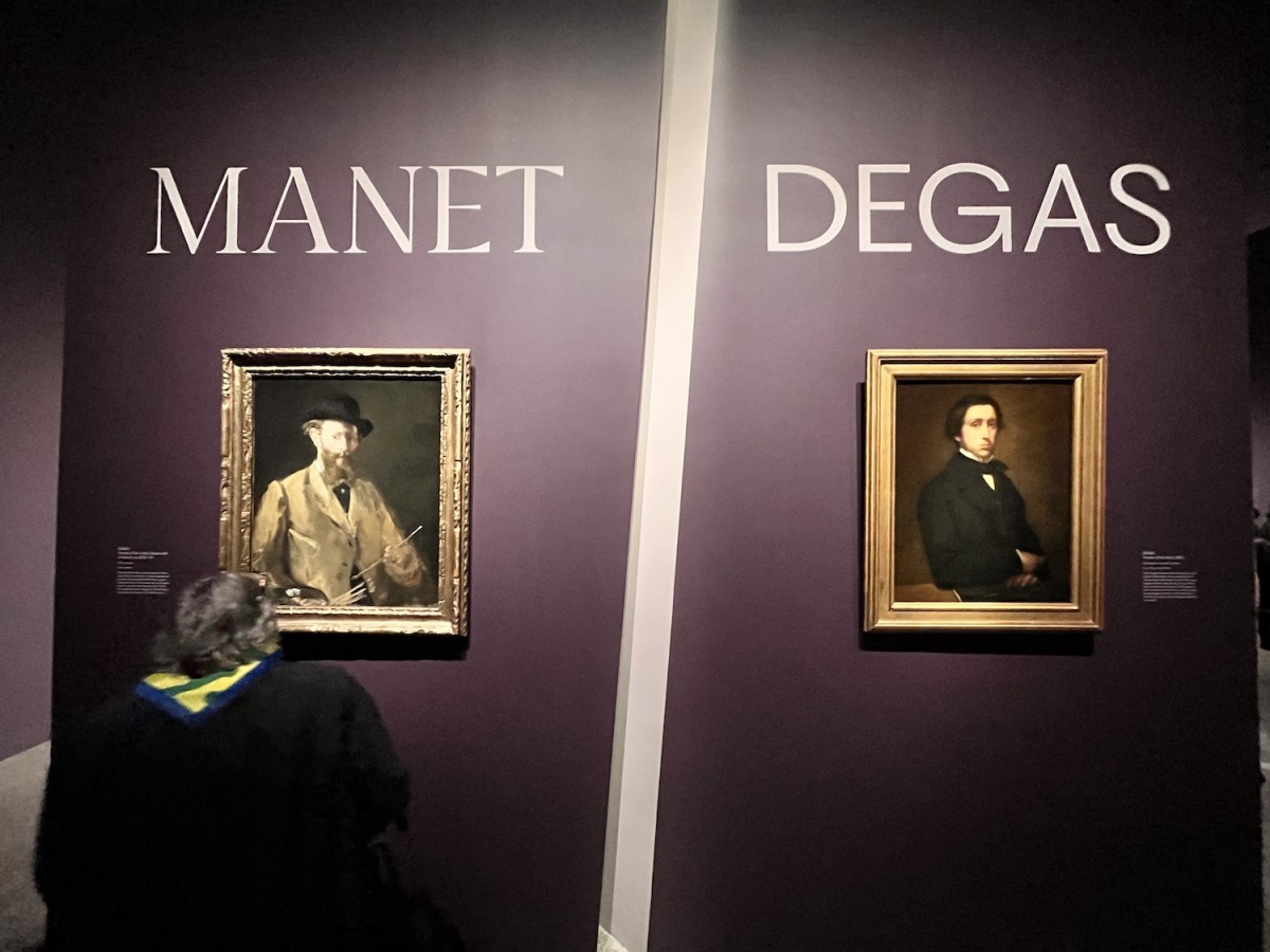 A purple wall, divided in the middle with a diagonal white line. On the left of the wall it says, “MANET,” and on the right it says, “DEGAS.” There is a gold-framed painting under each name. There is a person looking at the paintings.