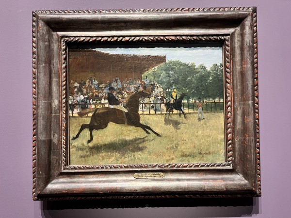 There is a framed painting on a light purple wall. The painting is of people on horses on a green field, with an audience in the background.