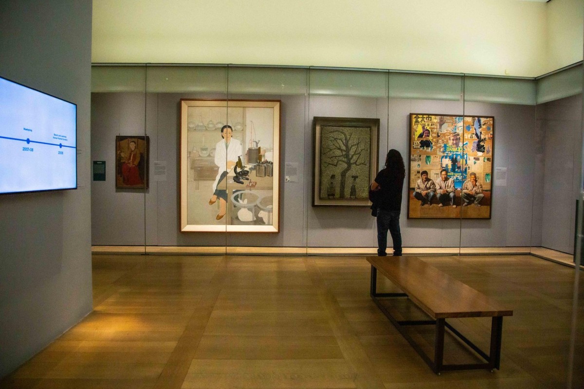 A room with wood flooring and gray walls. There are various paintings hung up behind glass screens.