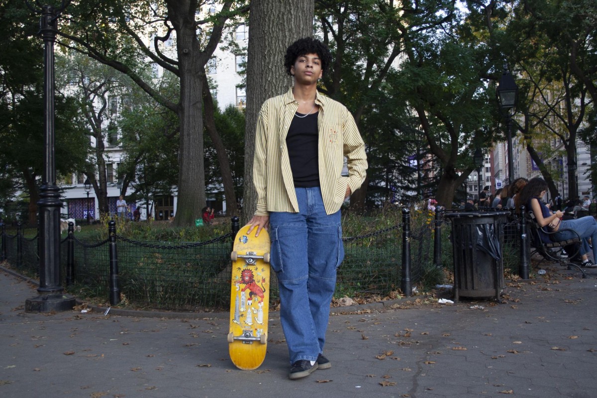 A person wearing blue jeans and a yellow button-up shirt stands in Washington Square Park holding a yellow skateboard with an image of a red lion.