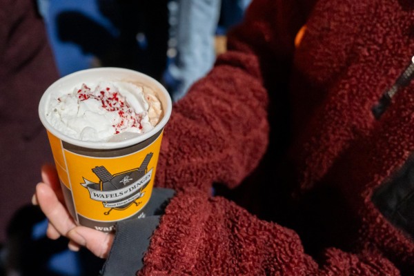 A person wearing a red jacket holds a yellow cup of hot chocolate with two hands. On top of the hot chocolate is a layer of whipped cream and red sprinkles.