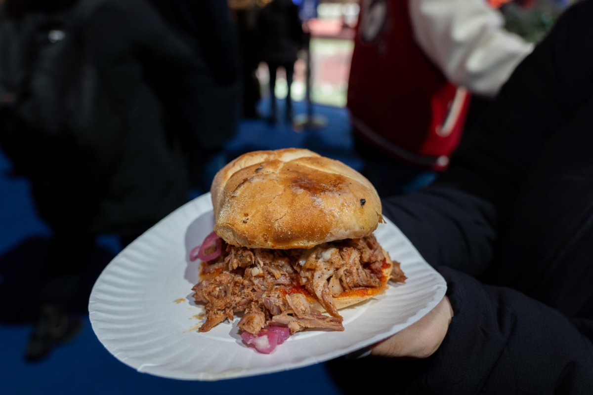 A person in a black coat holds out a pulled pork sandwich on a white paper plate.